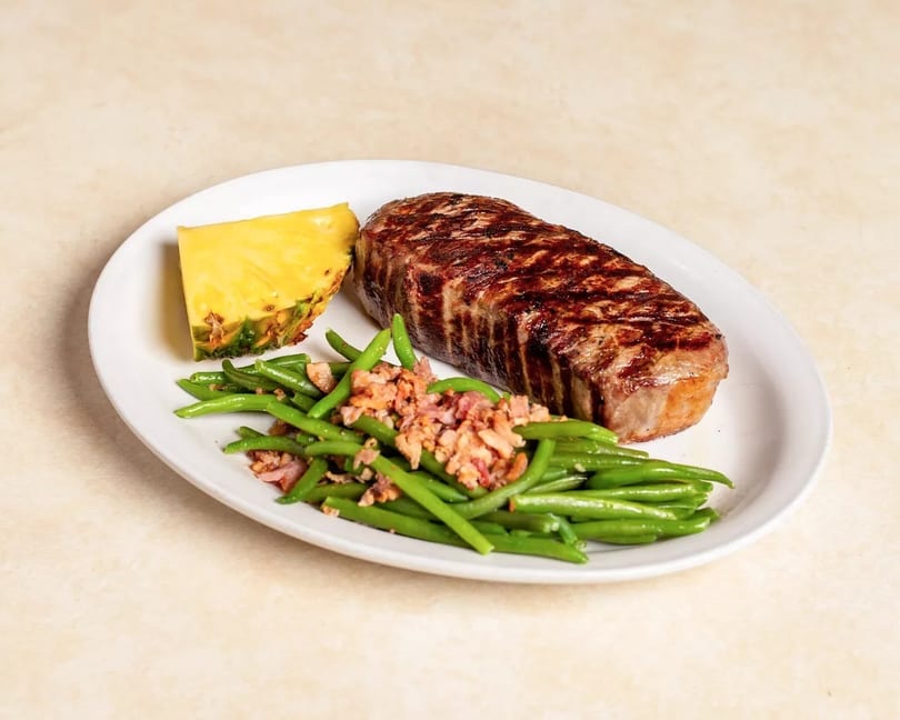 Grilled steak, green beans with bacon bits, and a pineapple wedge on a white plate.